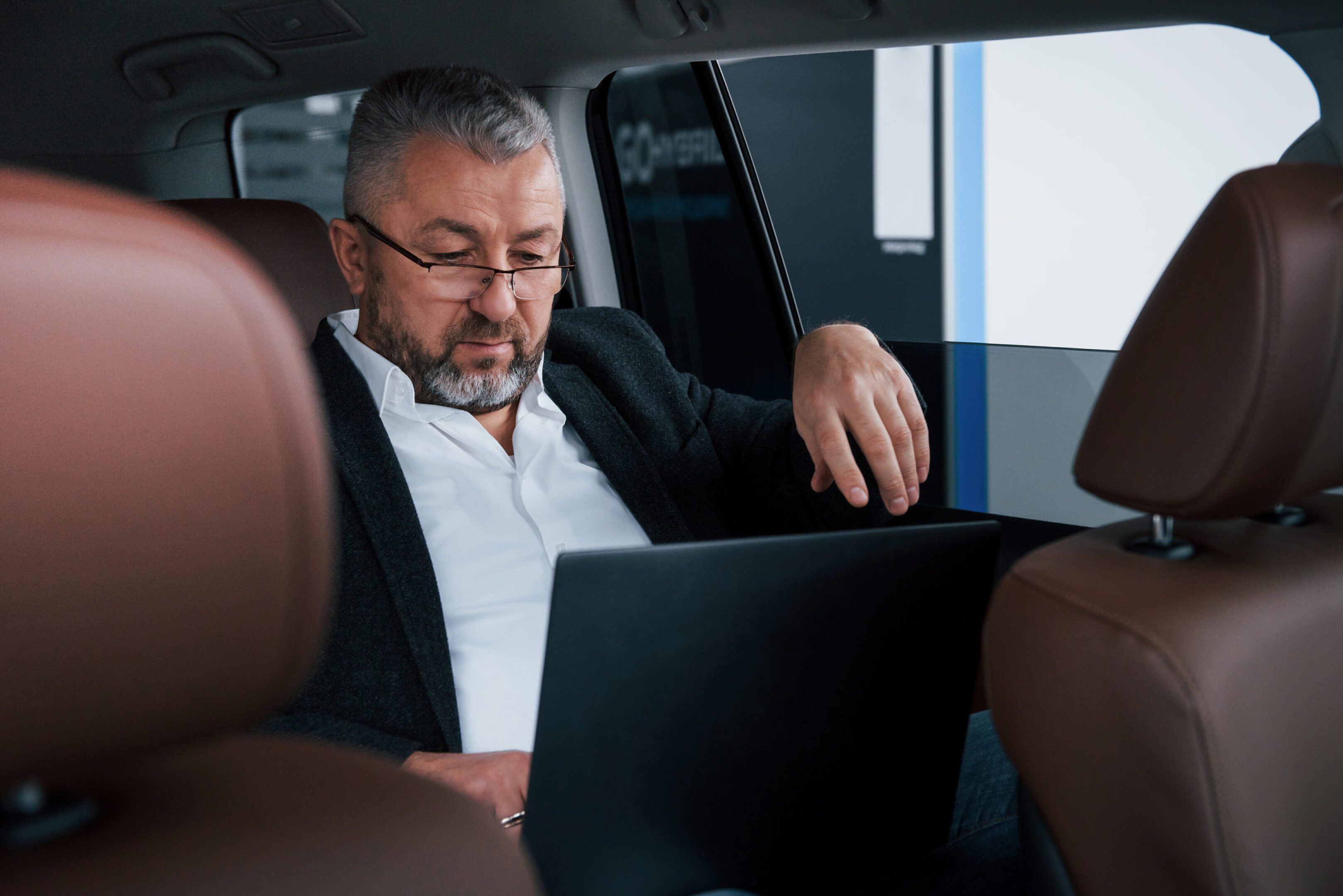 Job out of the office. Working on a back of car using silver colored laptop. Senior businessman A man sitting in the back of a car with his laptop.